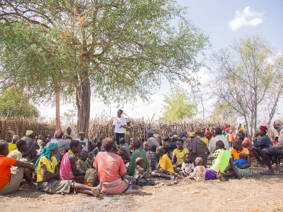 Villagers seated under a large tree for an outdoor community meeting